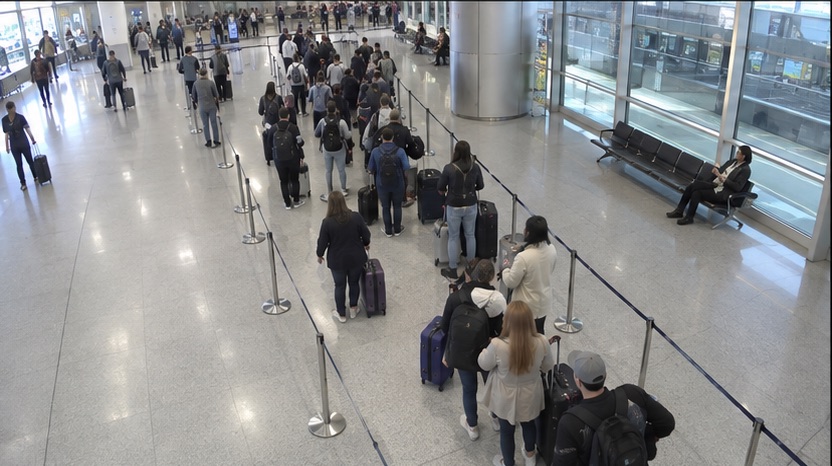 High-angle airport camera view showing passengers waiting with luggage