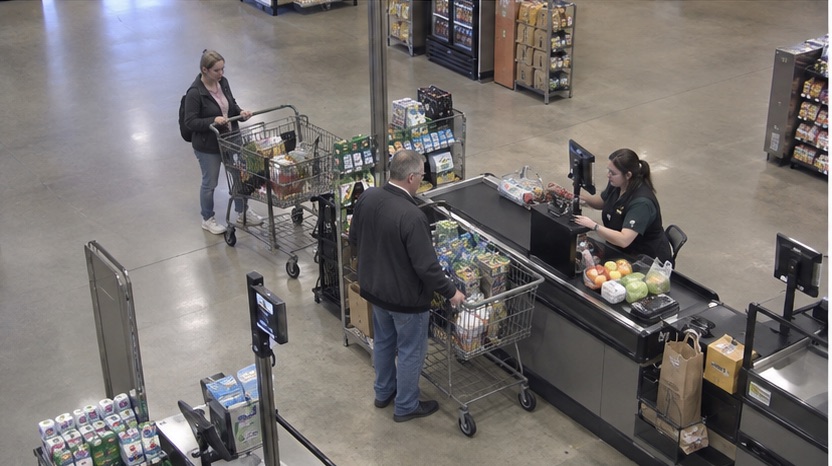 High-angle checkout camera view showing a transaction and the next waiting shopper